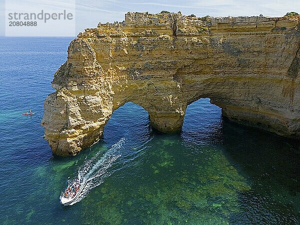 A large boat passes under natural arches made of rocks in crystal clear water on the coast  aerial view  Praia da Marinha  double rock arch  Lagoa  Rocky Algarve  Algarve  Portugal  Europe