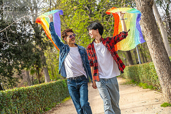 Happy multi-ethnic young gay couple raising LGBT rainbow colored fans in a park