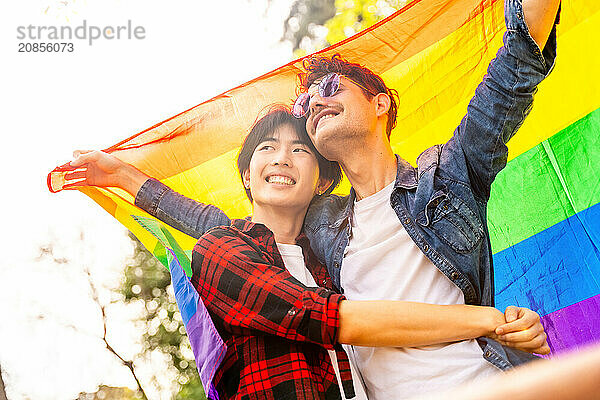 Low angle view of a multi-ethnic gay couple celebrating diversity raising lgbt flag in a park