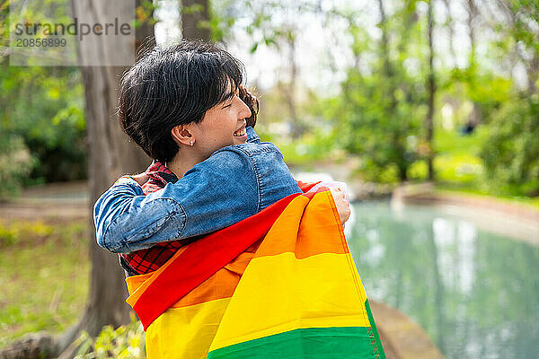 Multiracial gay couple embracing inside a lgbt flag standing in a park