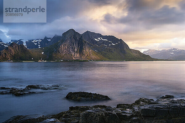 Landscape with sea and mountains on the Lofoten Islands  view across the fjord to the small town of Flakstad and the mountain Flakstadtinden as well as other mountains. Rocks in the foreground. At night at the time of the midnight sun in good weather. Some clouds in the sky  some sunlight on the mountains. Long exposure  blurred clouds. Early summer. Flakstadoya  Lofoten  Norway  Europe