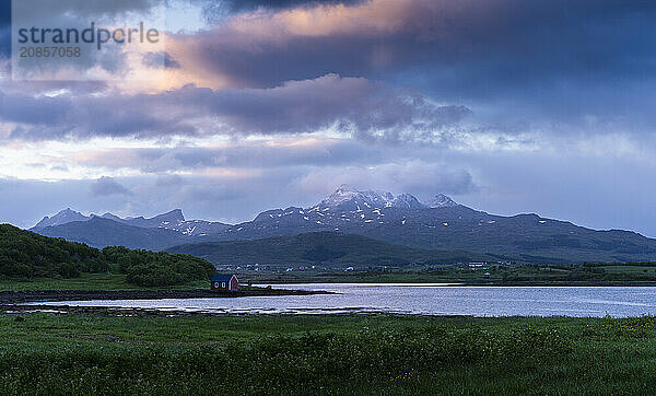 Landscape on the Lofoten Islands with meadows  sea and a small red hut  with other houses and mountains in the background. At night at the time of the midnight sun. Some coloured clouds in the sky. Lofoten  Norway  Europe