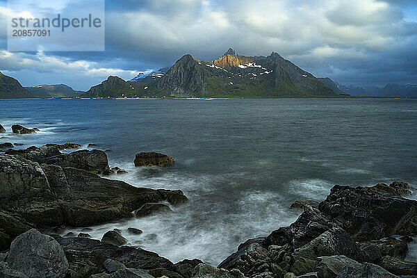 Landscape with sea and mountains on the Lofoten Islands  view across the fjord to the small town of Flakstad and the mountain Flakstadtinden as well as other mountains. Rocks in the foreground. At night at the time of the midnight sun  some clouds in the sky. Long exposure  blurred clouds  sun on a mountain peak. Early summer. Flakstadoya  Lofoten  Norway  Europe