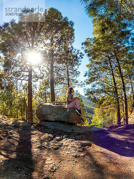 A woman sits on a rock in a forest. The sun is shining on her  creating a warm and peaceful atmosphere