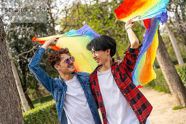 Multiracial gay couple raising lgbt rainbow hand fan smiling walking in a park