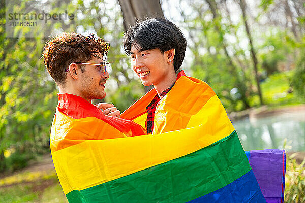 Multi-ethnic gay couple embraced in a park wrapped in rainbow flag