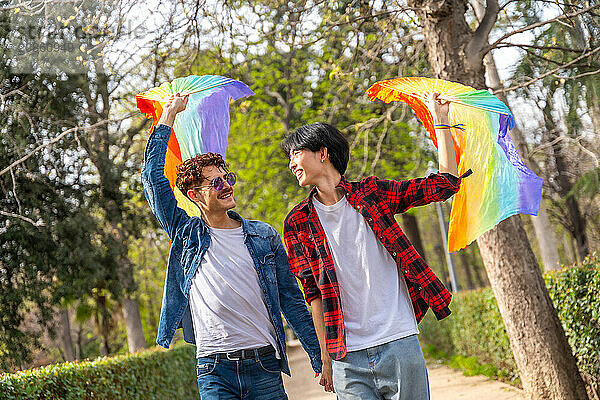 Happy multiracial gay couple walking holding hands and waving LGBT rainbow hand fans in a park