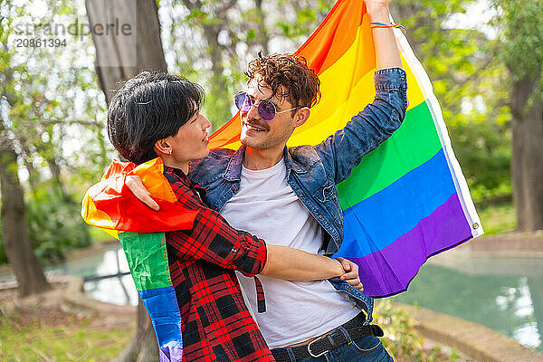 Happy and romantic multi-ethnic gay couple embracing wrapped in rainbow flag in a park