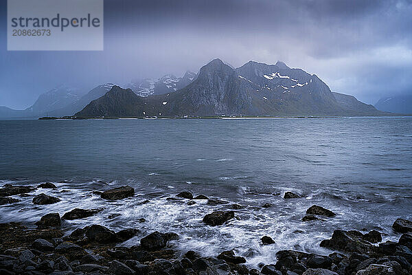 Landscape with sea and mountains on the Lofoten Islands  view across the fjord to the small town of Flakstad and the mountain Flakstadtinden as well as other mountains. Rocks in the foreground. At night at the time of the midnight sun  clouds in the sky  bad weather. Long exposure  early summer. Flakstadoya  Lofoten  Norway  Europe