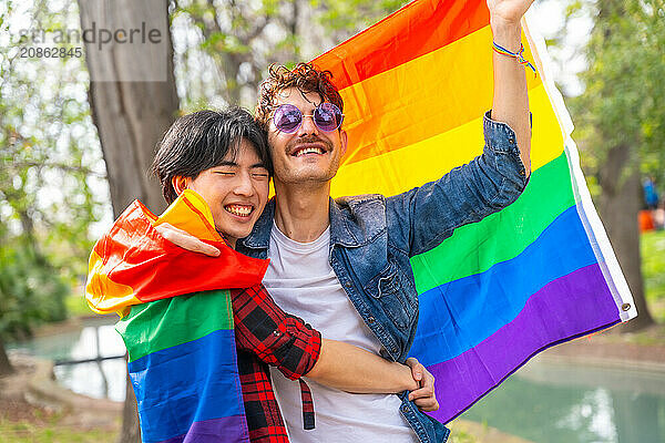 Happy multi-ethnic gay couple celebrating love waving lgbt flag in a park