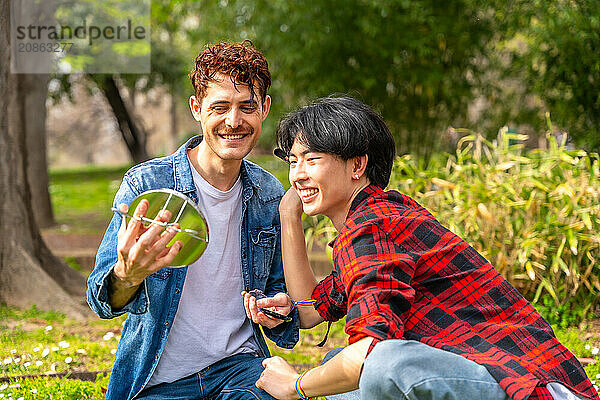 Gay couple making up using hand mirror sitting together in a park