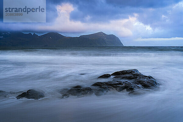 On the beach at Vikten. The sea washes around some rocks. Mountains in the background. At night at the time of the midnight sun  cloudy sky. Early summer. Long exposure. Vikten  Flakstadoya  Lofoten  Norway  Europe