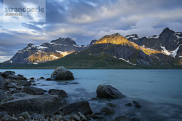 Landscape with sea and mountains on the Lofoten Islands  view over the fjord Flakstadpollen to mountains. Rocks in the foreground. At night at the time of the midnight sun  some clouds in the sky  sun spots on the mountains. Long exposure. Early summer. Flakstadoya  Lofoten  Norway  Europe