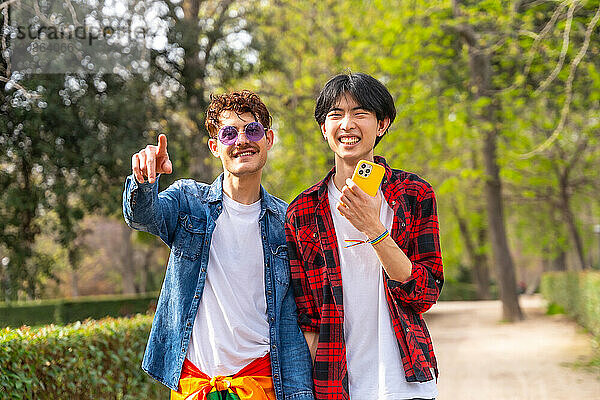 Multi-ethnic smiling gay couple standing pointing ahead using phone in a park