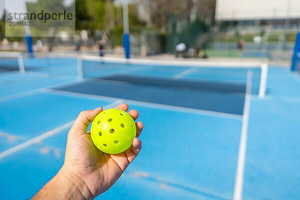 Unrecognizable person holding a plastic yellow pickleball ball with blue outdoor court on the background