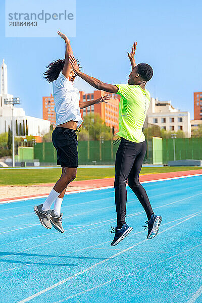 Excited running team of two african american young men jumping high-fiving in a running track