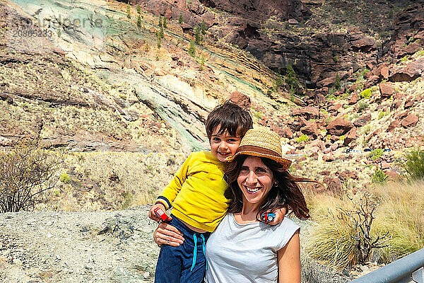 A woman and a child are posing for a picture in front of a rocky mountain. The woman is wearing a straw hat and the child is wearing a yellow shirt. Scene is lighthearted and joyful  as the mother