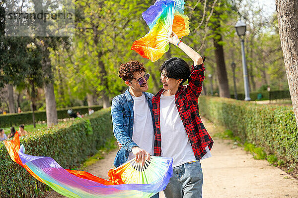 Multiracial gay couple waving lgbt rainbow fans while walking together along a public park