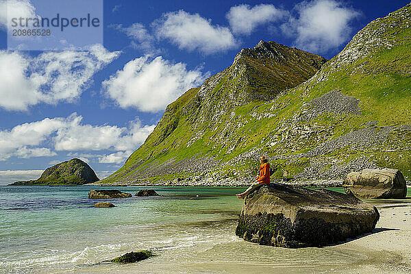 Landscape with sea at the sandy beach of Haukland (Hauklandstranda) with the mountain Veggen. A woman sits barefoot on a rock and looks out to sea. Shot during the day with blue sky  some clouds. Early summer. Haukland Beach  Haukland  Vestvagoya  Lofoten  Norway  Europe