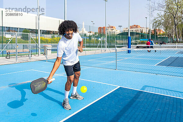 Full length photo of an smiling man with afro hair serving while playing pickleball in an outdoor court