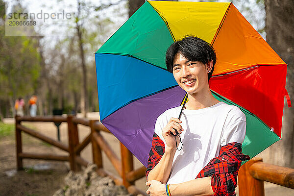 Asian gay man posing with rainbow umbrella standing in a park smiling at camera