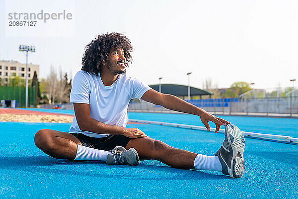 Side view of a smiling young african sportive man stretching sitting on a running track