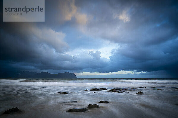 On the beach at Vikten. The sea washes around some rocks. Mountains in the background. At night at the time of the midnight sun. Cloudy  dramatic sky. Early summer. Long exposure. Vikten  Flakstadoya  Lofoten  Norway  Europe