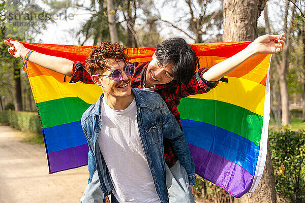 Portrait of a joyful multi-ethnic gay couple waving lgbt flag piggybacking in a park