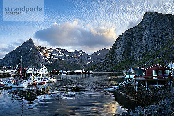 The village of Hamnoy. On the right  a typical red wooden house (rorbuer) on wooden stilts. On the left  other houses and boats in a small harbour. In the background the mountain Olstinden. At night at the time of the midnight sun in good weather  some clouds in the sky. Early summer. Hamnoy  Moskenesoya  Lofoten  Norway  Europe