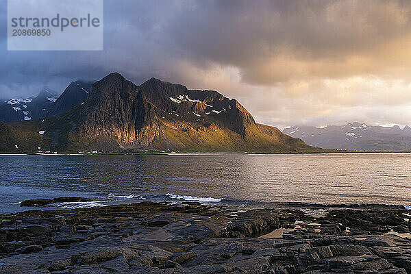 Landscape with sea and mountains on the Lofoten Islands  view across the fjord to the small town of Flakstad and the mountain Flakstadtinden as well as other mountains. Rocks in the foreground. At night at the time of the midnight sun. A few clouds in the sky  the sun shines sideways on the mountains. Golden hour. Early summer. Flakstadoya  Lofoten  Norway  Europe