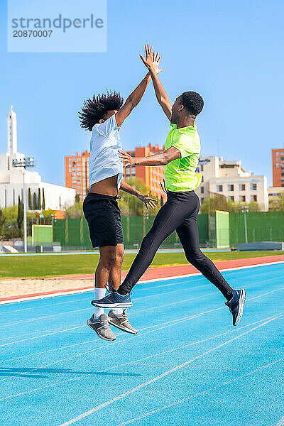 Vertical photo of two african american young friends jumping celebrating and clasping hands in a running track