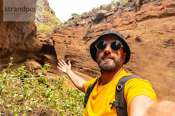 A man in a yellow shirt and sunglasses is taking a selfie in front of a rocky cliff. Concept of adventure and exploration  as the man is posing in a natural setting with a backpack on