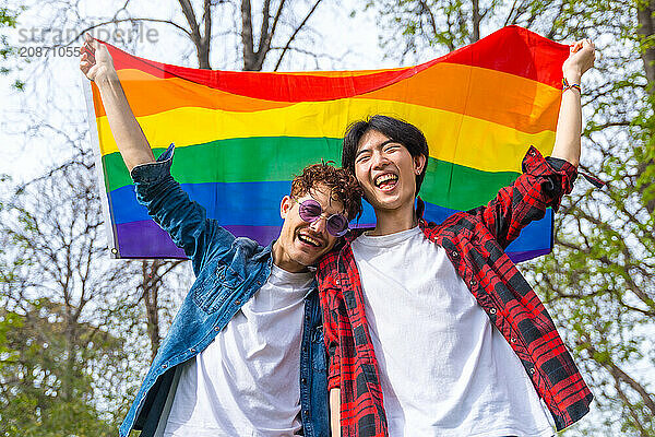 Low angle view portrait of a happy gay couple laughing while raising lgbt rainbow flag outdoors in a park