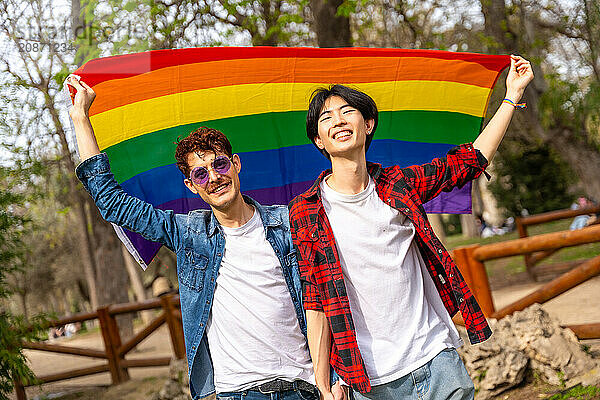 Portrait of a multi-ethnic gay couple raising a rainbow lgbt flag in a park