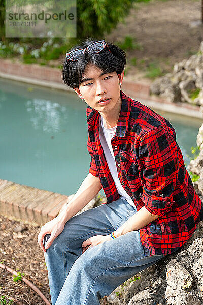 Vertical photo of an Asian gay man sitting next to urban stream of water in a park