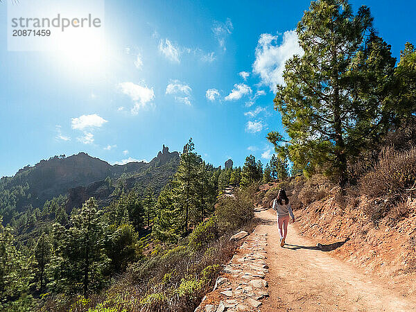 A woman is walking on a dirt path in a forest. The sky is blue and the sun is shining