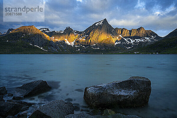 Landscape with sea and mountains on the Lofoten Islands  view over the fjord Flakstadpollen to the mountain Stortinden and others. A rock in the foreground. At night at the time of the midnight sun  some clouds in the sky  the sun shining on the mountains. Long exposure. Early summer. Flakstadoya  Lofoten  Norway  Europe