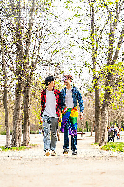 Vertical full length photo of a gay male couple walking along a park carrying LGBT rainbow flag