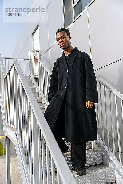 Vertical portrait of an African beauty man posing with long black jacket in stairs