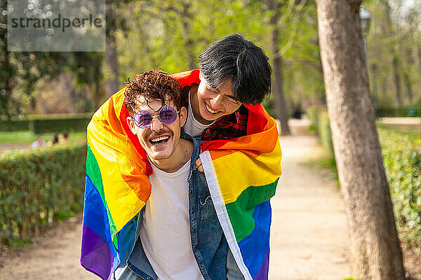 Multi-ethnic gay men wrapping in lgbt flag piggybacking in a park