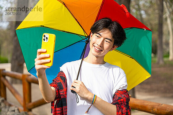 Chinese gay man taking a selfie with mobile phone while holding a rainbow colored umbrella standing in a park