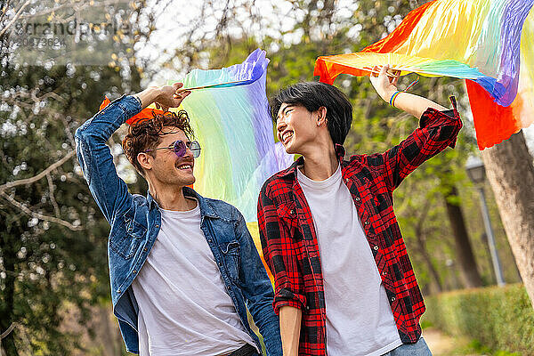 Gay couple holding hands strolling happily in a park raising lgbt rainbow hand fans