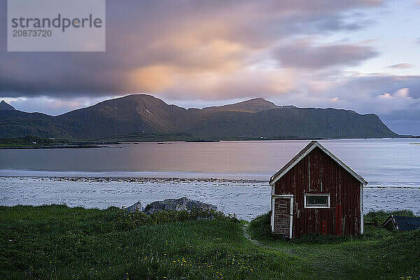 A red wooden hut on the sandy beach of Ramberg (Rambergstranda)  with the sea behind it. At night at the time of the midnight sun. Clouds in the sky  the sun shines on clouds and mountains. Early summer. Long exposure  blurred clouds. Ramberg  Flakstadoya  Lofoten  Norway  Europe