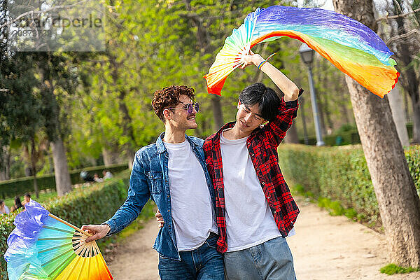 Multiracial gay couple celebrating love waving lgbt rainbow fans in a park