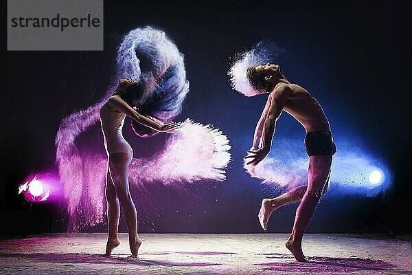 Young sporty man and slim woman posing emotionally in jump in cloud of pink and blue dust studio portrait