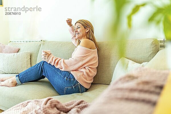 Horizontal photo full length and copy space of a blonde casual adult woman celebrating raising fist sitting relaxed on the sofa