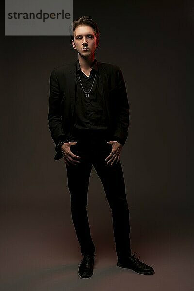 Full body of confident young male model in black leather jacket and pants looking at camera against black background