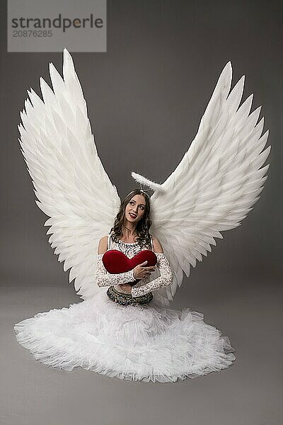 Young female sitting in studio on gray background wearing white dress angel wings hold red heart