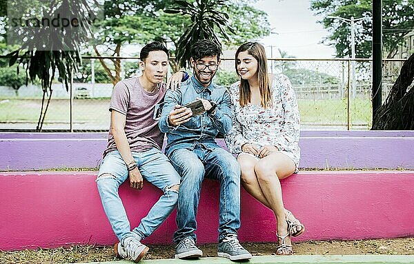 Three happy friends looking at the cell phone sitting in the park. Three happy people looking at cell phone outdoors