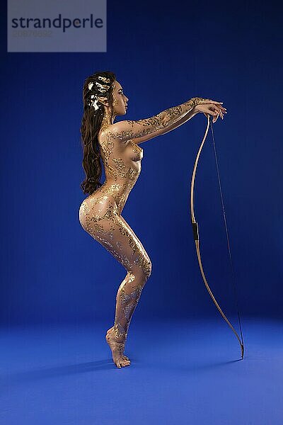 Full length of sensual confident female model with golden paint on naked body and elf ears with arrows and bow dance on blue background